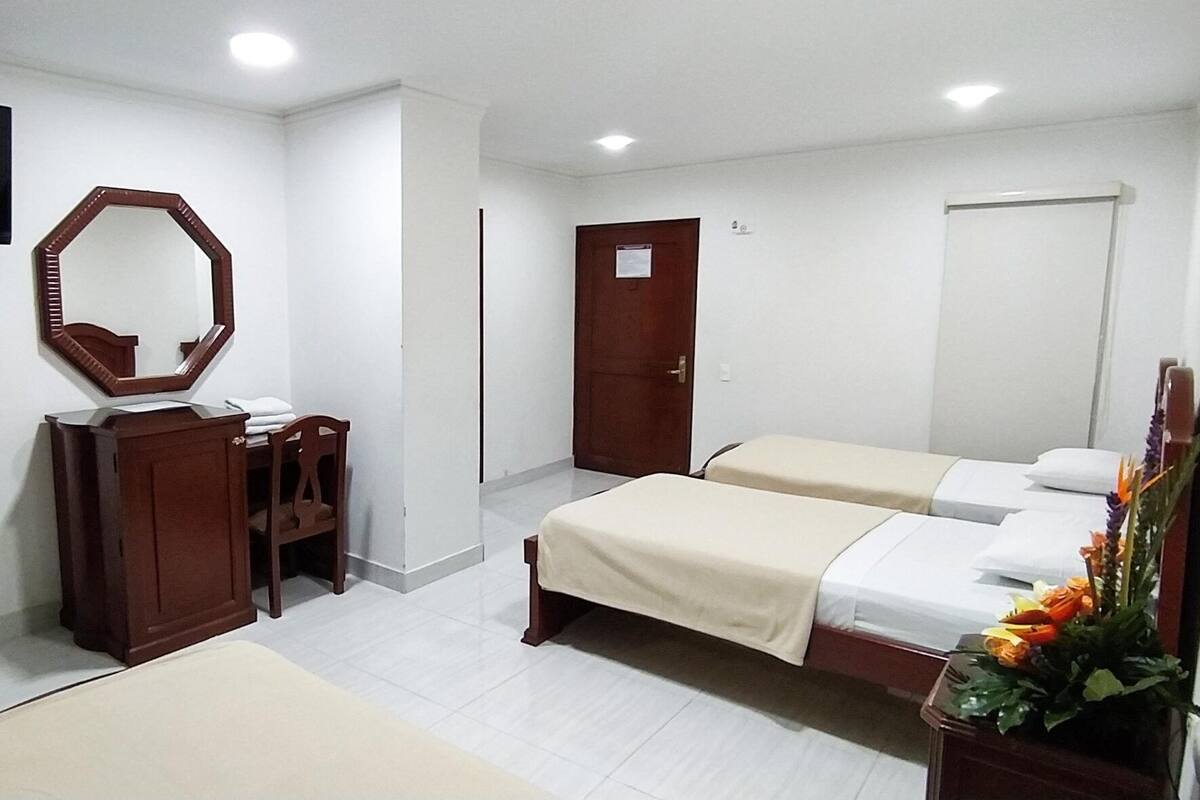 Habitación de hotel con televisión, cómoda de madera, espejo, cama con cubrecama color beige y una puerta.