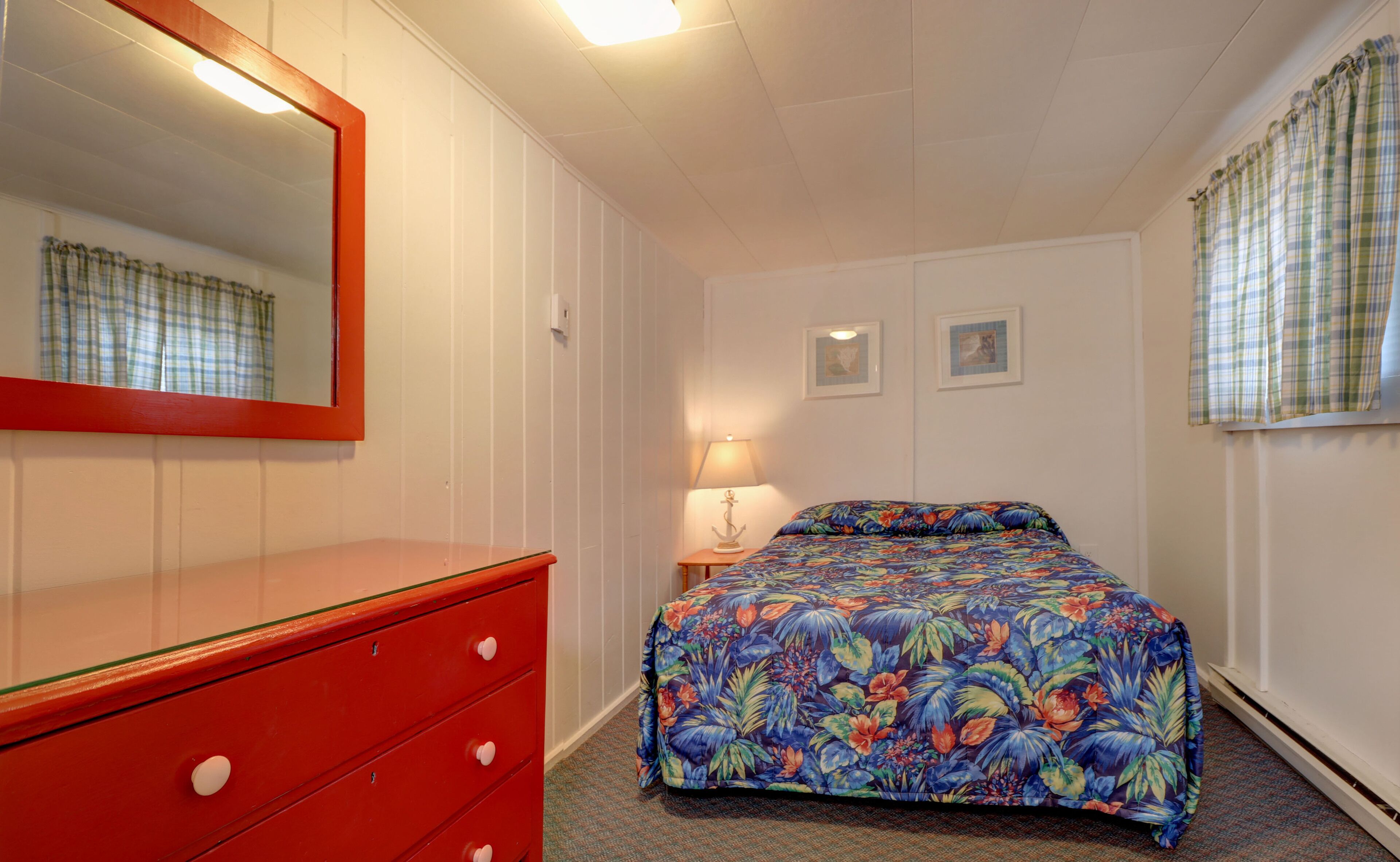 A bedroom with a red dresser, a floral bedspread, a mirror, and two framed pictures on the wall.