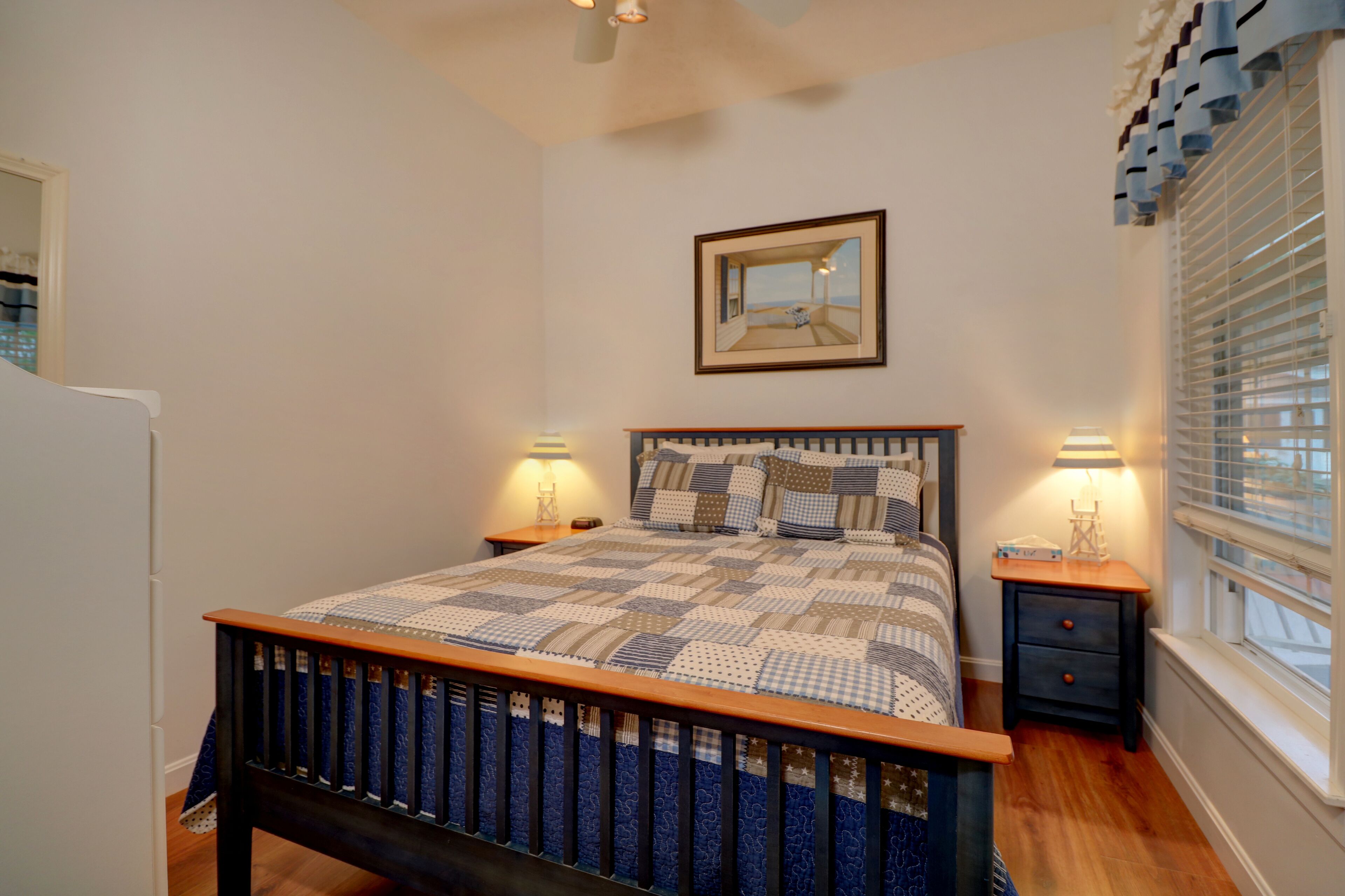 A bedroom with a wooden bed frame, a quilted bedspread, two bedside tables with lamps, a mirror on the wall, and a window with blinds.