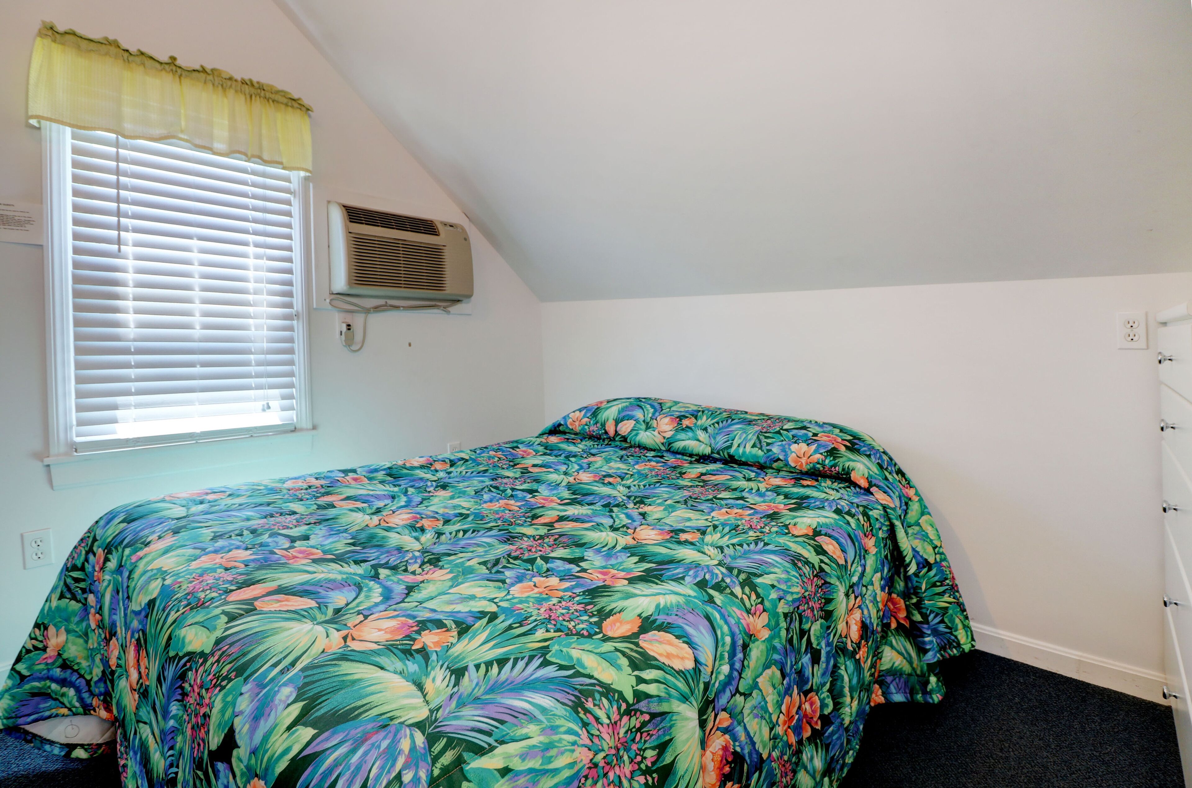 Un dormitorio con cama, ventana con persianas, equipo de aire acondicionado y toma de corriente en la pared.