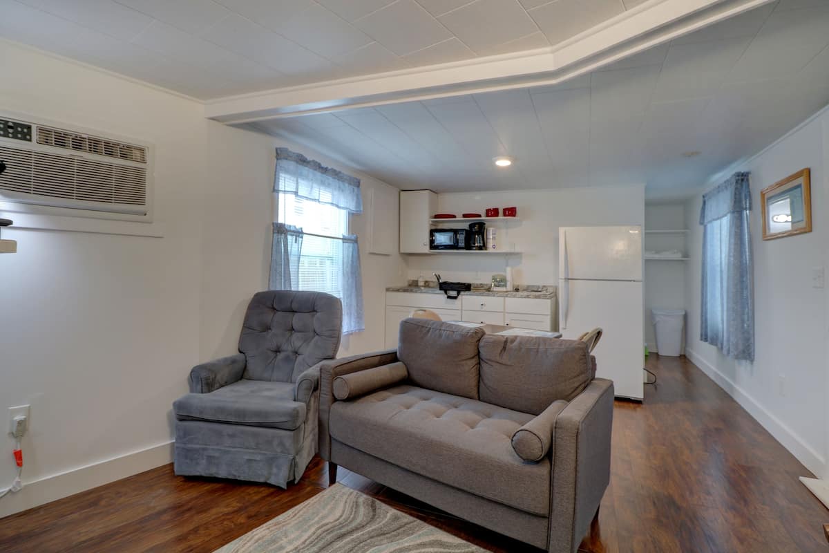 Een woonkamer met een grijze fauteuil, een grijze bank en een houten vloer. Er is een keukenhoek met een magnetron, een koffiezetapparaat en een koelkast.