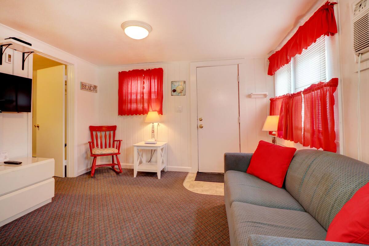 Een kamer met een grijze bank, rode kussens, een rode stoel, een witte tafel en een televisie aan de muur.