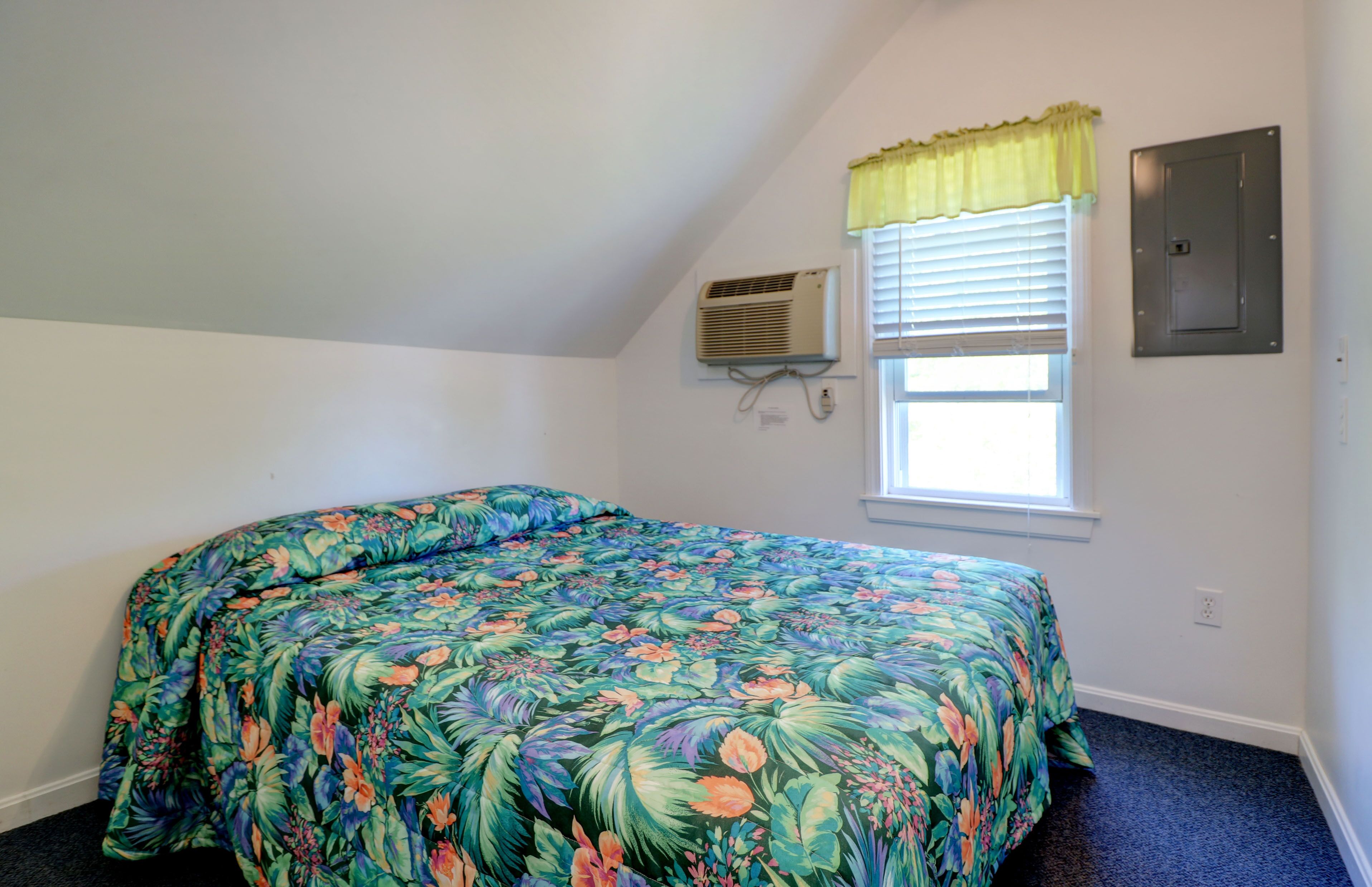 A bedroom with a floral bedspread, a window with blinds, an air conditioning unit, and a wall-mounted electrical panel.