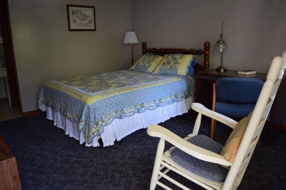 A bedroom with a bed, a chair, a nightstand, a lamp, and a framed picture on the wall.