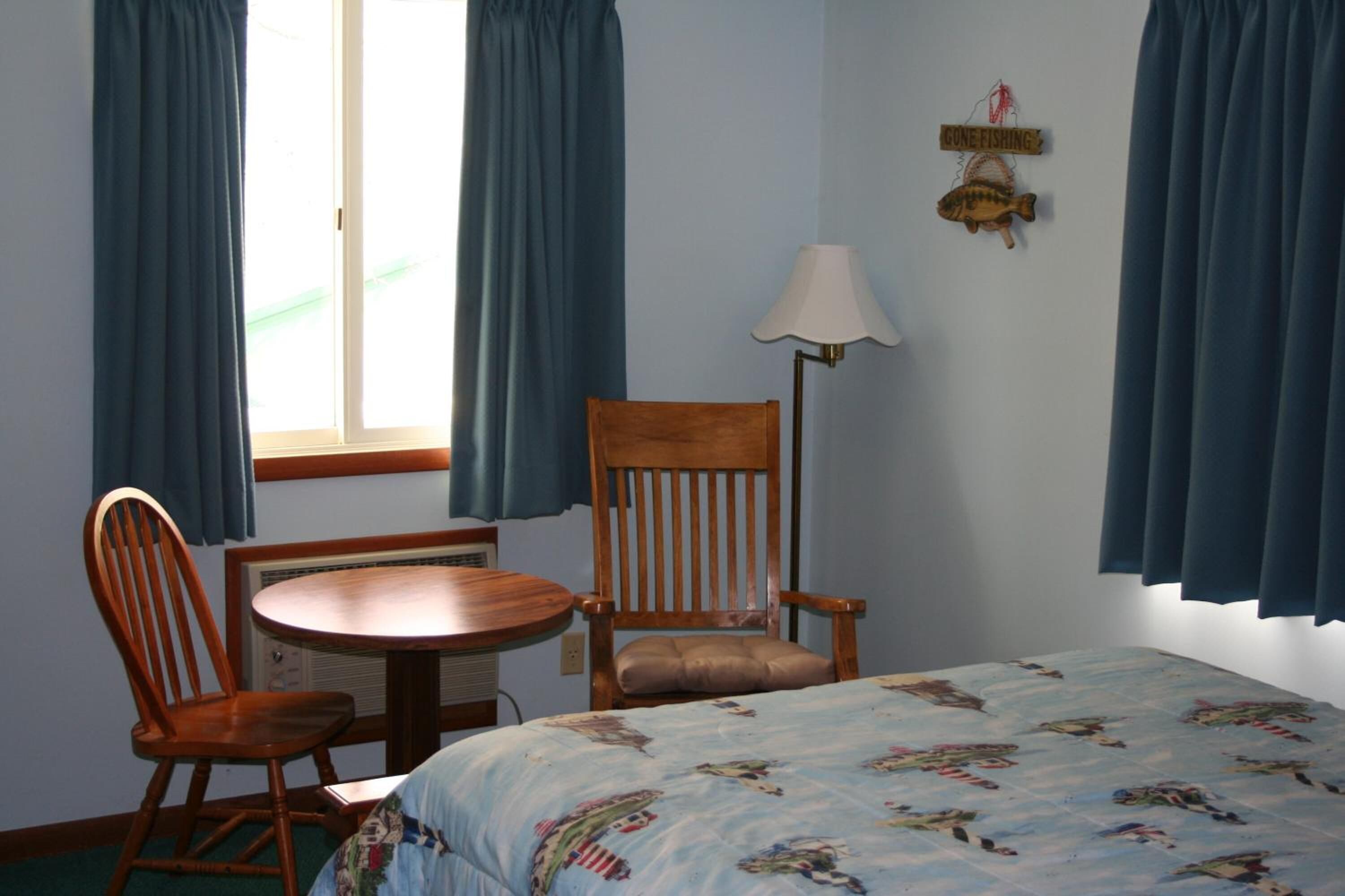 A room with a bed, a wooden chair, a table, and a lamp.