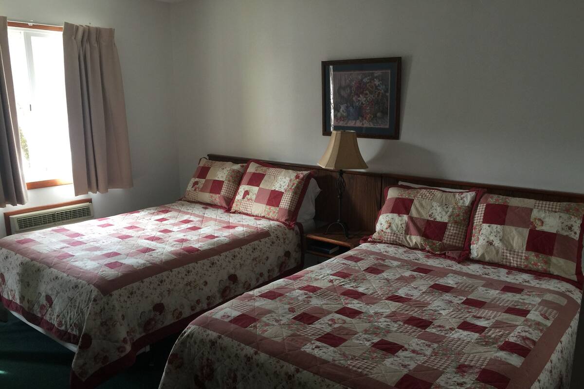 A room with two beds, each with floral-patterned quilts, a standing lamp, and a framed picture on the wall.