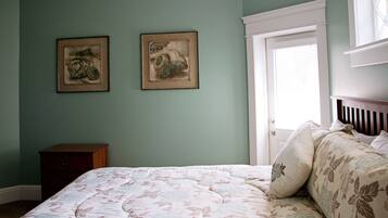 A bedroom with a bed, two framed pictures on the wall, a nightstand, and a window with blinds.