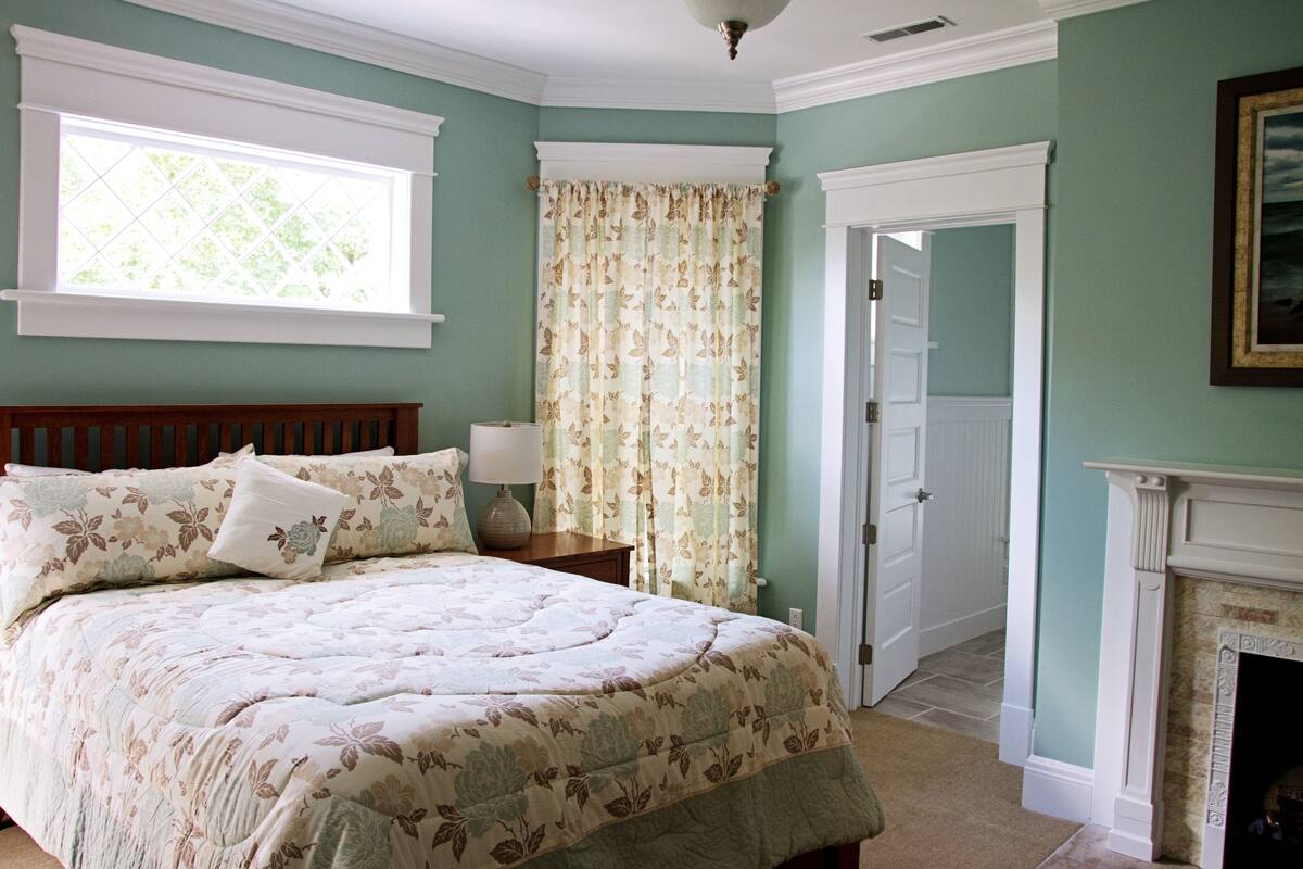 A bedroom with a bed, a nightstand, a window, a door, and a fireplace.