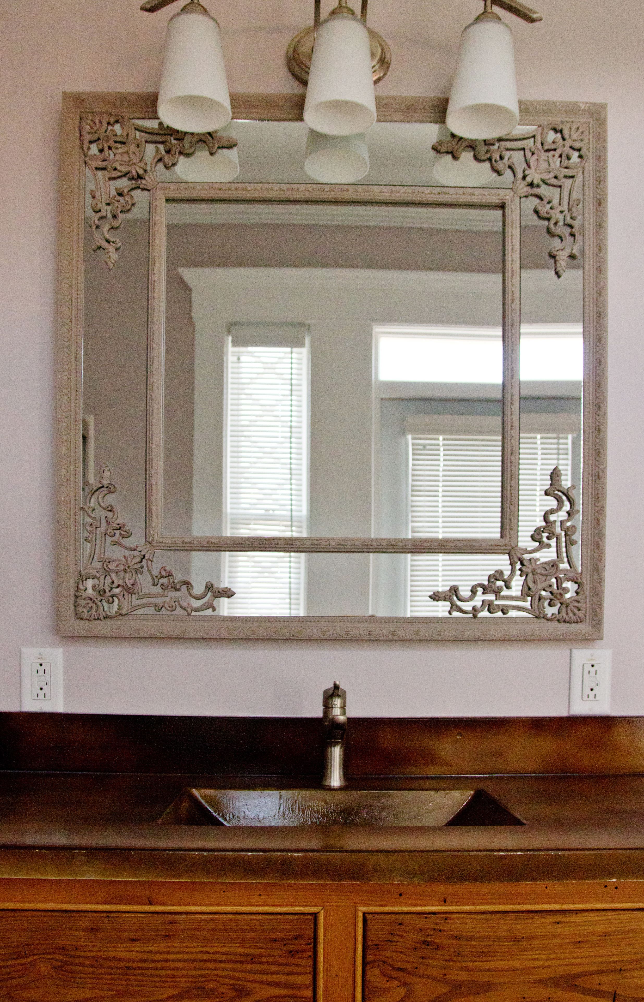 A bathroom with a large ornate mirror, three wall-mounted lights, and a wooden vanity.