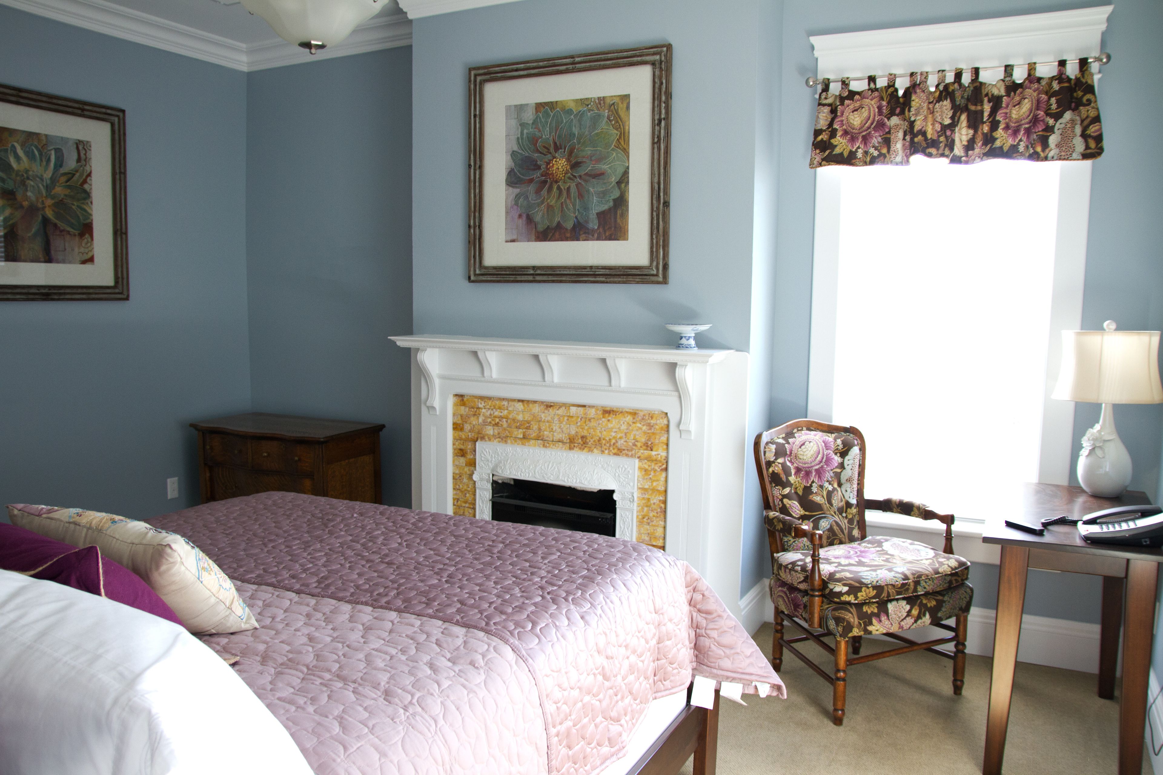 A bedroom with a bed, a chair, a fireplace, and a desk with a lamp.