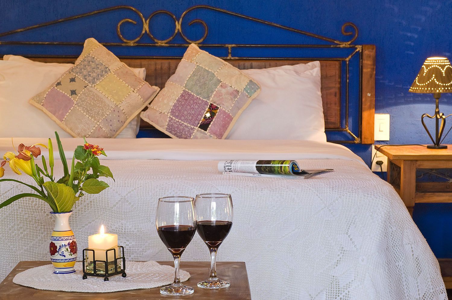 Una cama bien hecha con una colcha blanca, dos almohadas decorativas, una mesita de noche con una botella y un vaso, y un florero con flores.