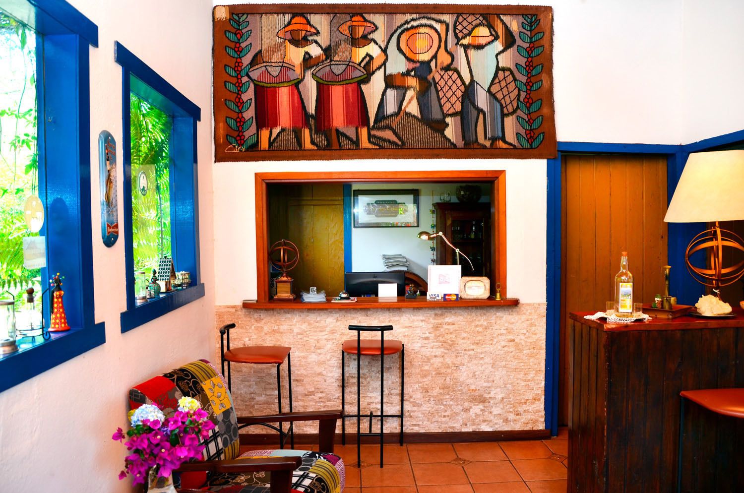 Una habitación con pared de ladrillo, una ventana con contraventanas azules, un cuadro sobre la ventana, dos sillas, una mesita con una lámpara y un florero con flores.