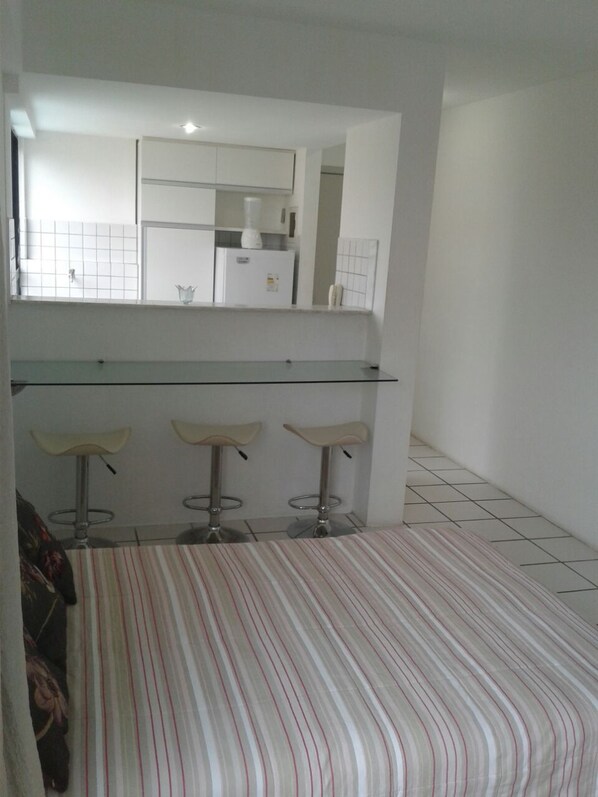 Una habitación con una cama a rayas, una zona de bar con superficie de vidrio y taburetes, y una cocina visible a través de un arco.
