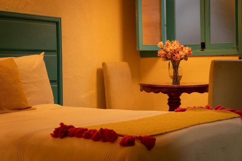 Une chambre d’hôtel avec un lit, une table de chevet ornée d’un vase de fleurs et une fenêtre avec des volets verts.
