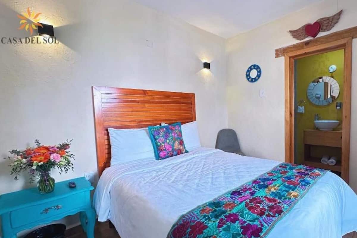 Une chambre à coucher avec une tête de lit en bois, un lit recouvert d’une literie blanche, une courtepointe aux couleurs vives, une table de chevet ornée d’un vase de fleurs et un luminaire fixé au mur.