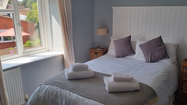 A neatly made bed with pillows, a bedside table with a lamp, and a window with curtains. - Winchester Arms (Taunton)