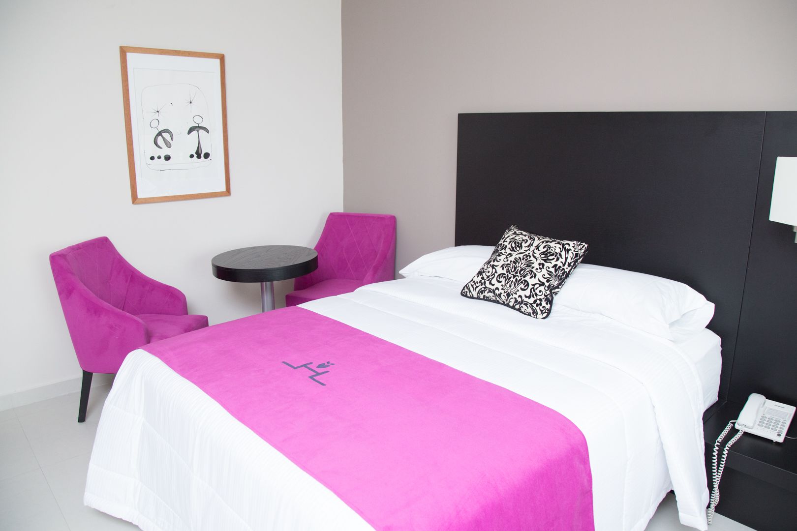 Habitación de hotel con cama, una mesa redonda, dos sillas, un cuadro en la pared y un teléfono en la mesita de noche.
