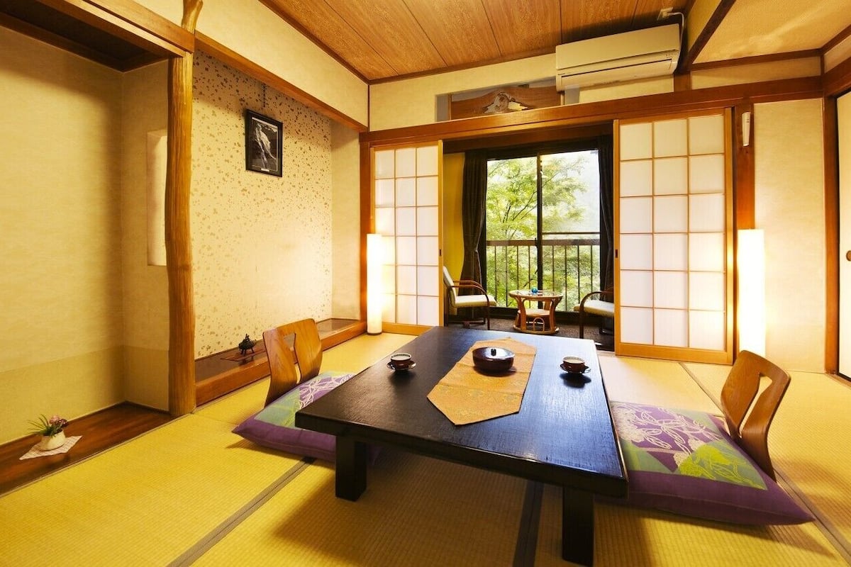 Une pièce de style japonais traditionnel, avec un sol recouvert de tatamis, une petite table sur laquelle sont disposés des ustensiles à thé, des portes coulissantes et une vue sur la verdure environnante.