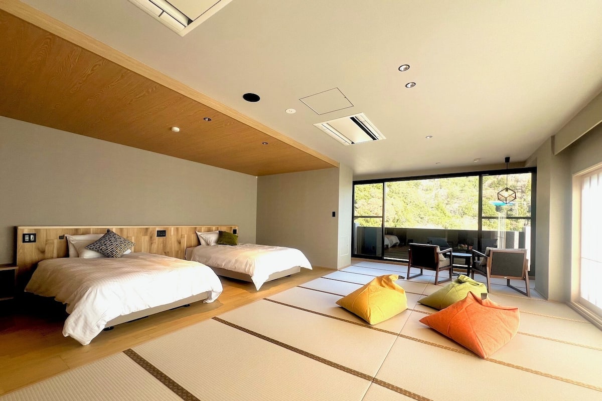 Une chambre d’hôtel moderne avec deux lits, des tatamis, un coin repas avec des chaises et de grandes fenêtres donnant sur l’extérieur.