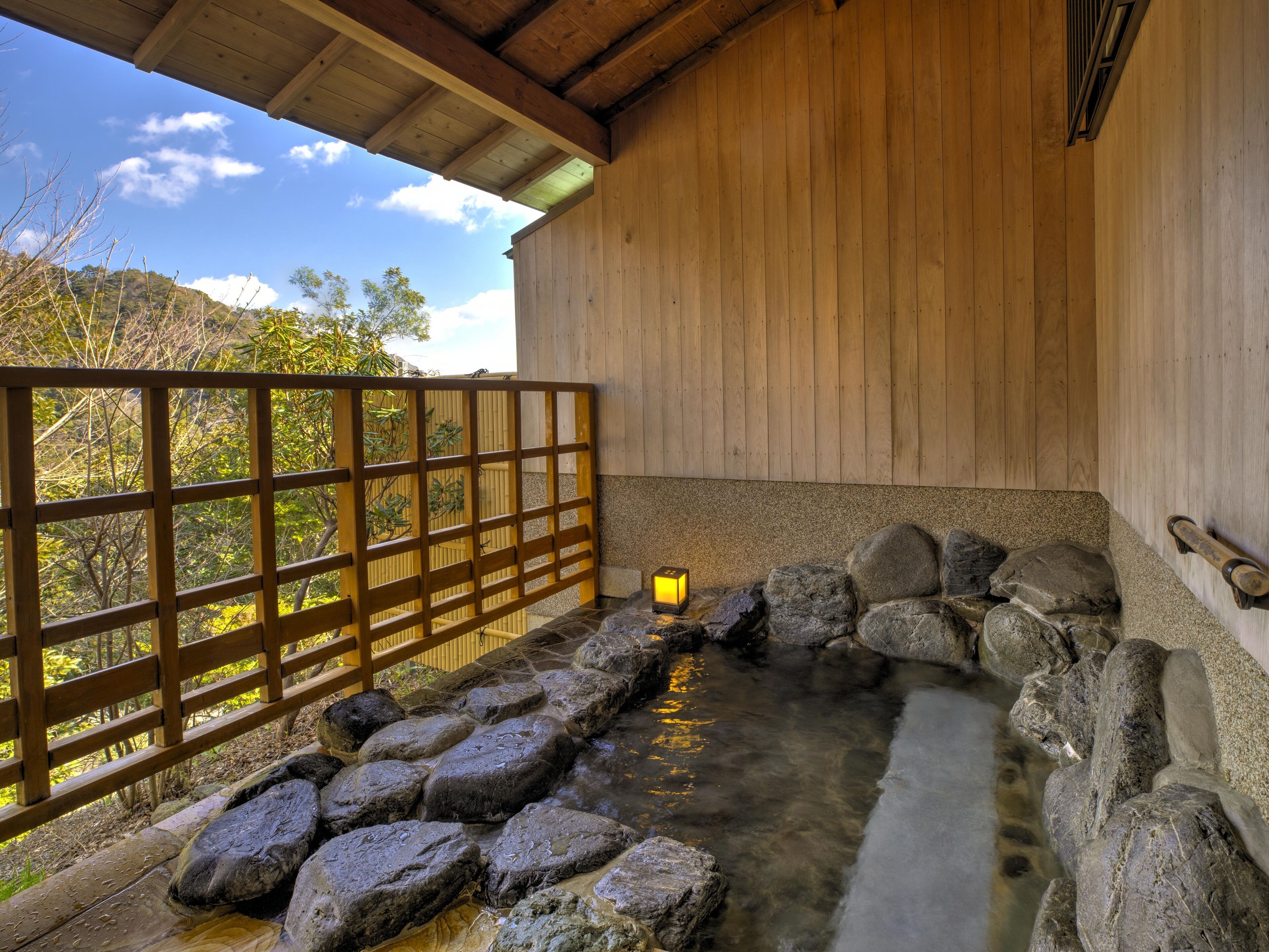 Un onsen japonés tradicional con interior de madera, manantial termal de piedra y valla de madera con vistas a un paisaje natural.