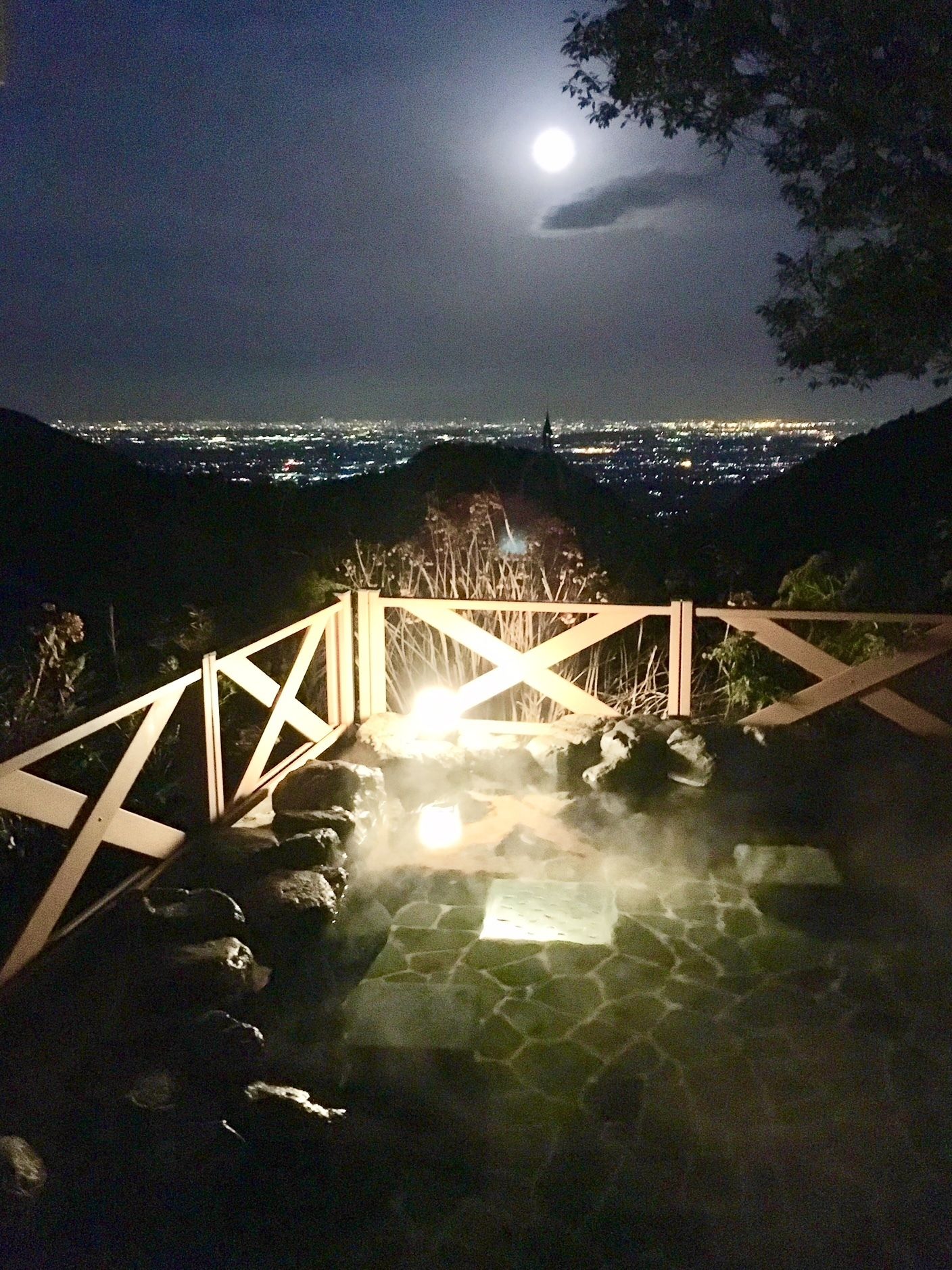 A nighttime view of a cityscape with a moon in the sky, a wooden staircase leading to a hot spring area with steam rising, and a railing overlooking the city.
