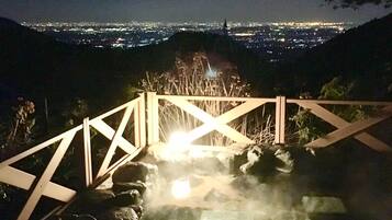 A nighttime view of a cityscape with a moon in the sky, a wooden staircase leading to a hot spring area with steam rising, and a railing overlooking the city.