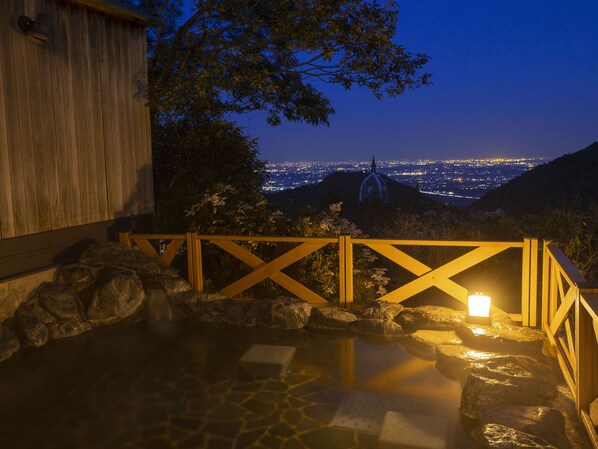 Un área de aguas termales con una terraza de madera y vista a un paisaje urbano nocturno.