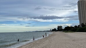 A wide sandy beach with a calm sea, a tall building on the right, and a partly cloudy sky. - Baan Thew Talay Blue Sapphire (Cha-am)