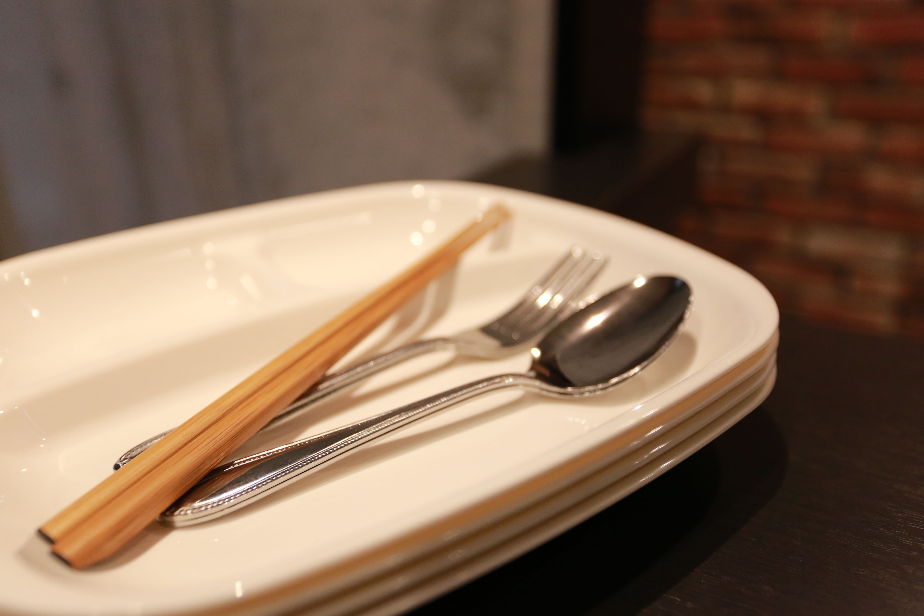 Une assiette blanche avec des baguettes, une fourchette et une cuillère, posée sur une surface sombre.