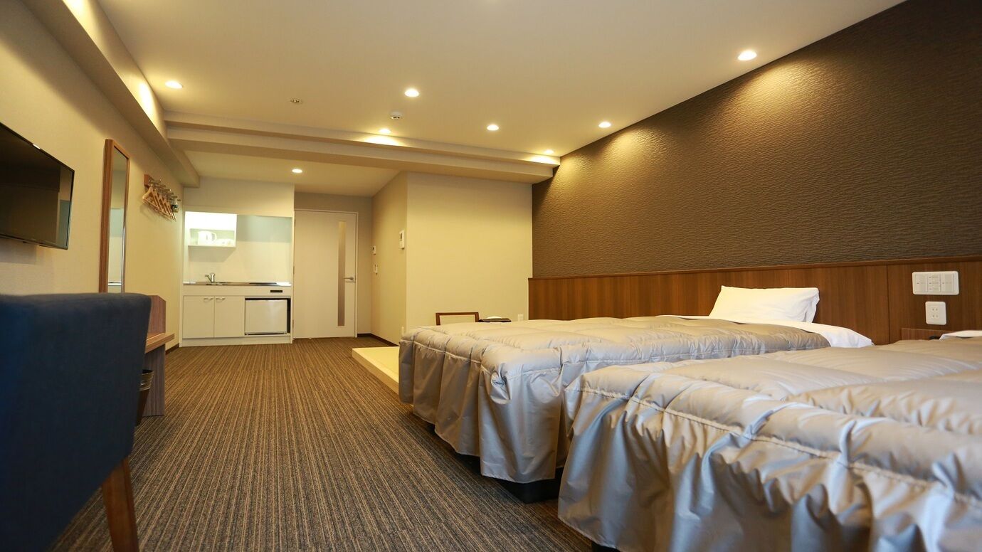 A hotel room with two beds, a desk, a chair, a TV, and a kitchenette.