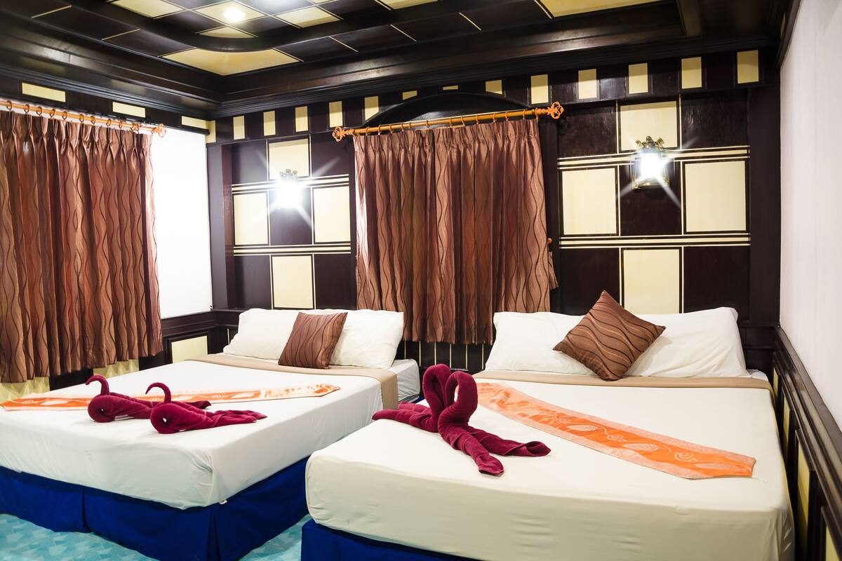 A hotel room with two beds, each with a white mattress and brown headboard. The room has a dark wooden ceiling with a geometric pattern and a window with curtains.