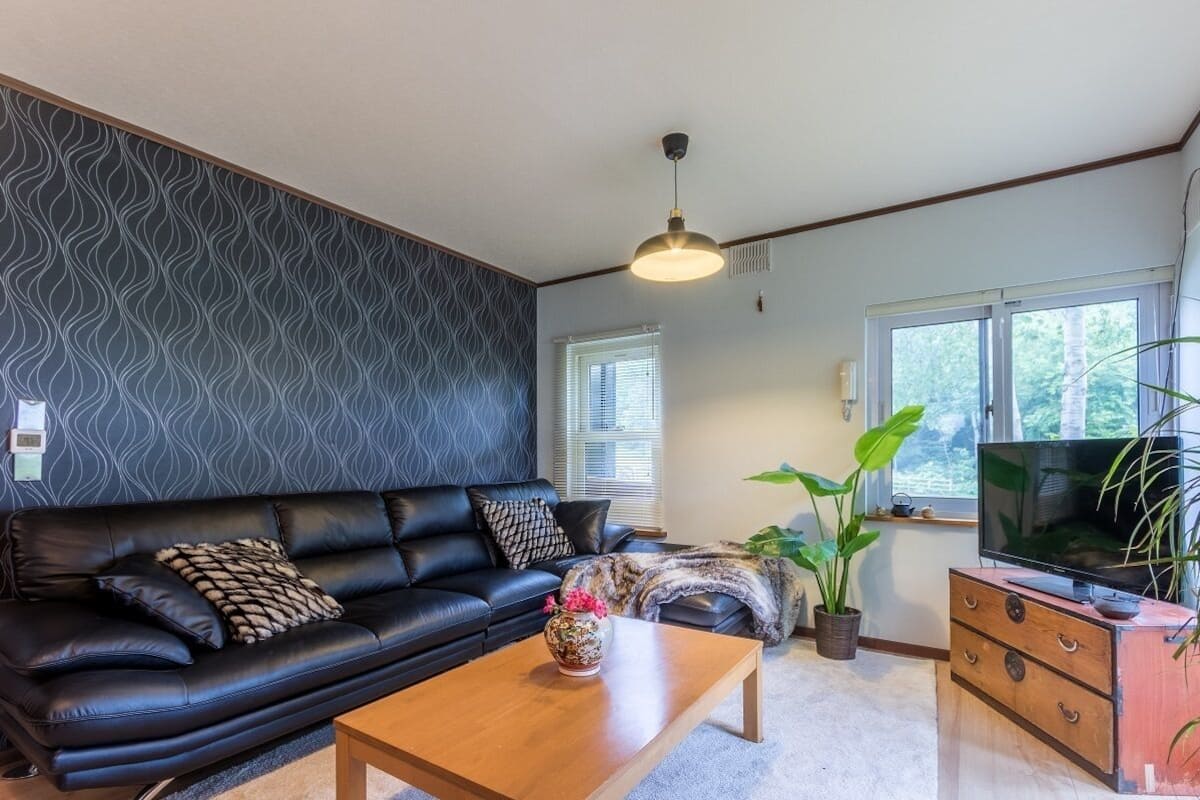 A living room with a black leather sofa, a wooden coffee table, a TV on a wooden cabinet, and a window with blinds.