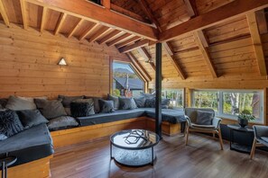 A log cabin interior with a large wooden beam ceiling, a built-in bench seating area, a round glass table, and a view of the outside through a window. - Hooting Owl Lodge (Niseko)