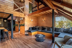 A spacious loft-style room with a wooden ceiling, a built-in seating area, a dining table with chairs, and a large window offering a view of the outdoors. - Hooting Owl Lodge (Niseko)