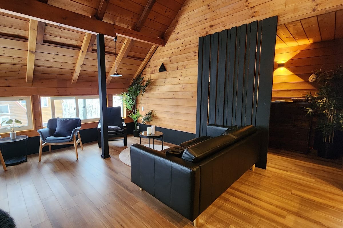 Un intérieur chaleureux avec des éléments en bois, un plafond haut, un canapé noir, un fauteuil gris et une table à manger en bois.