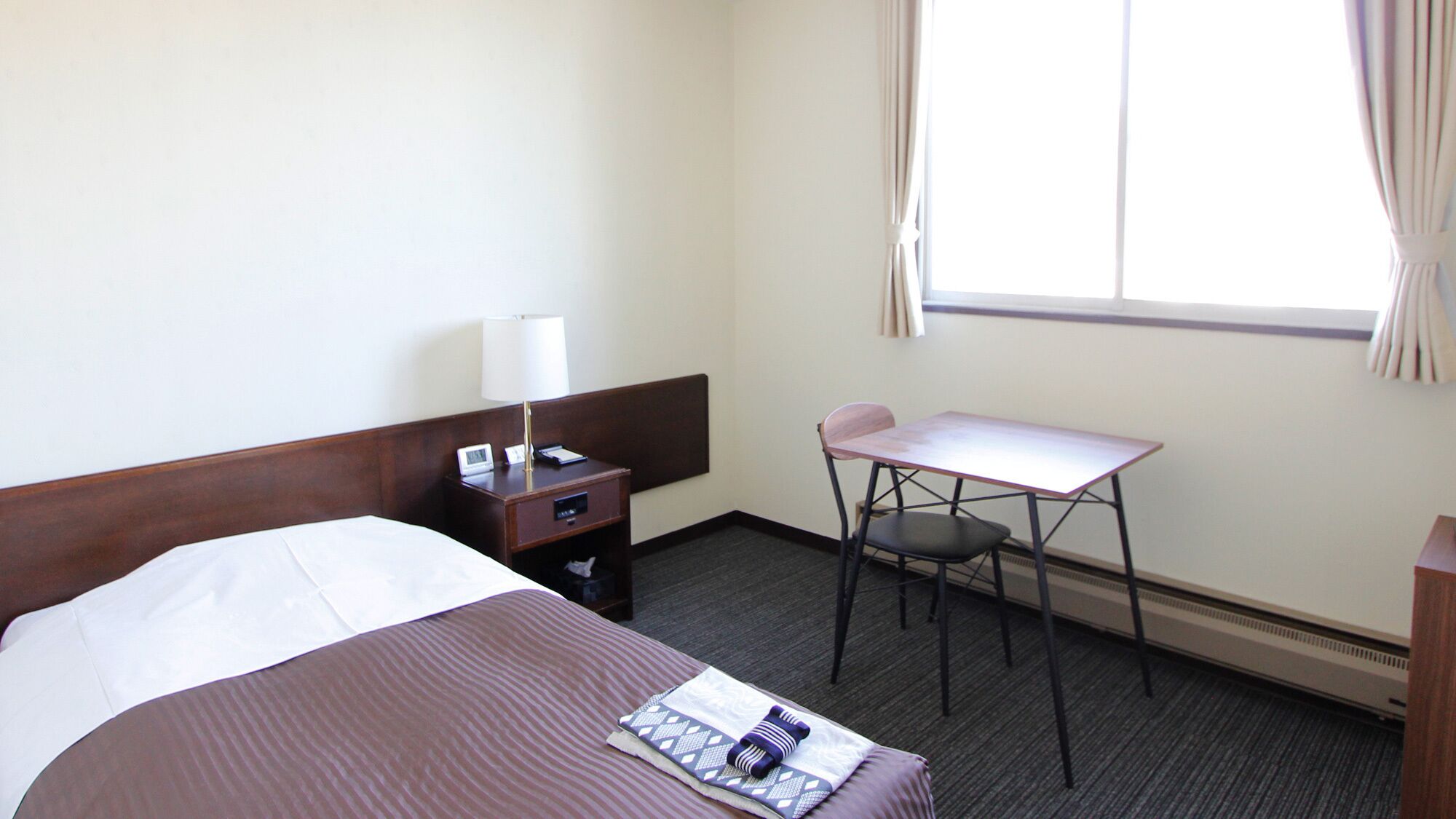 A hotel room with a bed, a desk, a chair, a lamp, and a window.