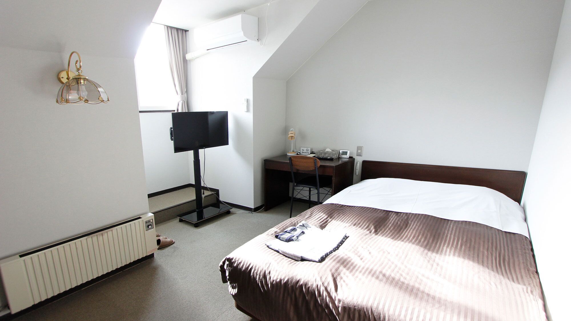 A hotel room with a bed, a desk with a chair, a television, and a window with curtains.