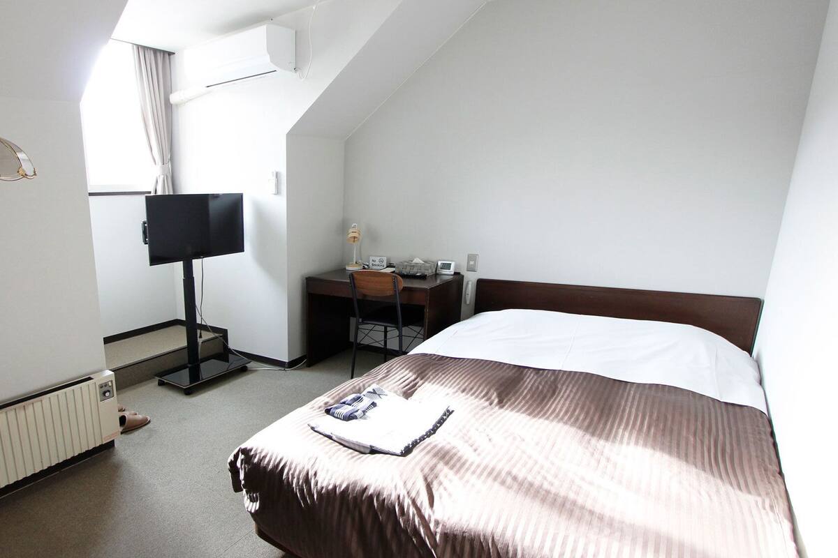 A hotel room with a bed, a desk with a chair, a television, and a window with curtains.