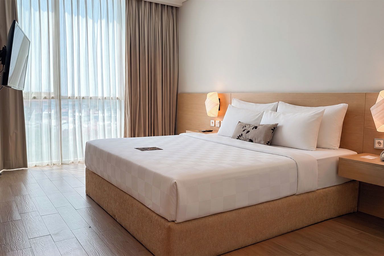 A hotel room with a large bed, a bedside lamp, a flat-screen TV mounted on the wall, and a window with curtains.