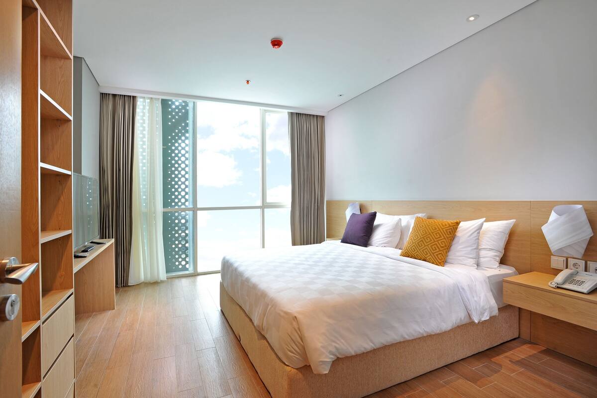A modern hotel room with a large bed, wooden shelving, a bedside table with a lamp, and a view of the sky through the window.