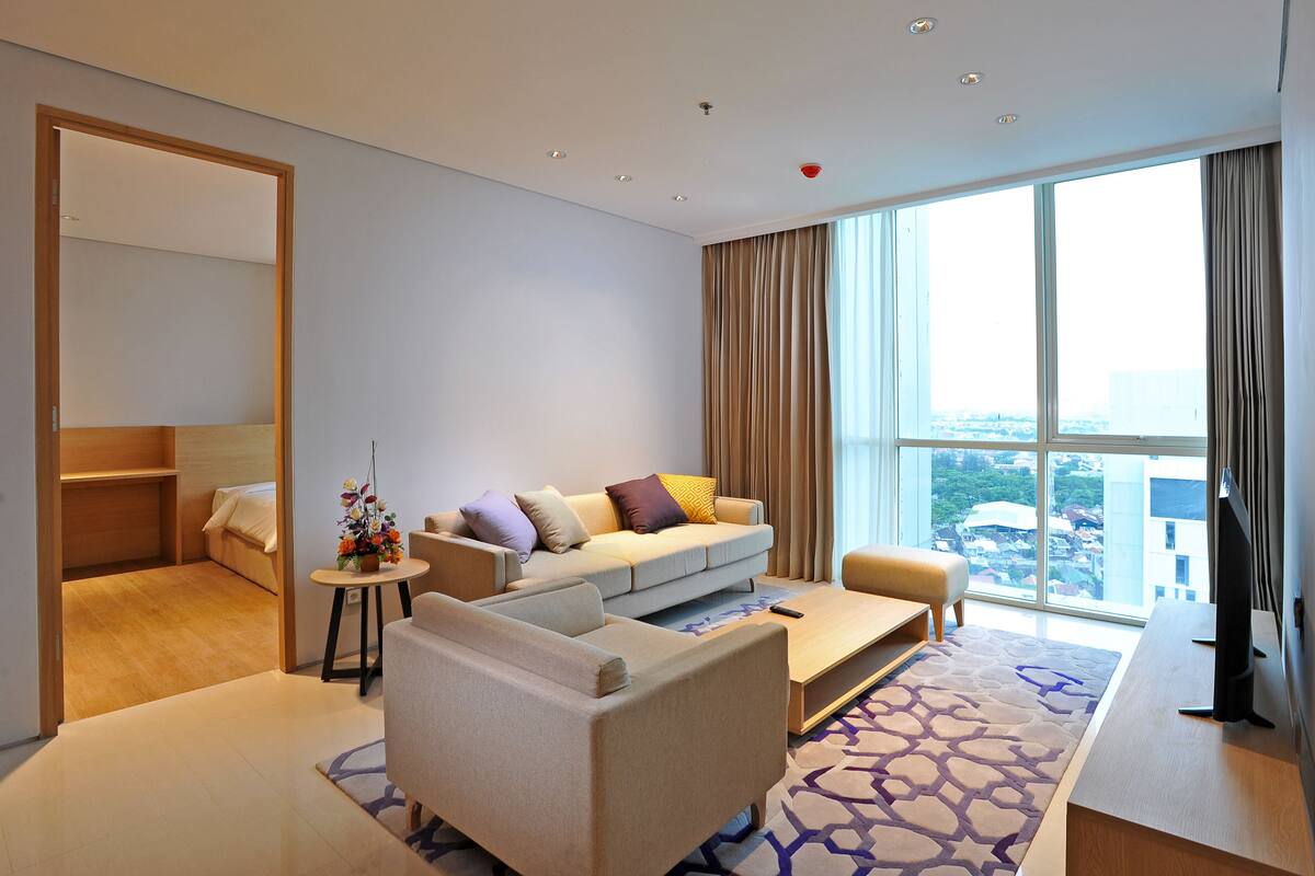A modern hotel room with a sofa, armchair, and a small table. There is a large window with curtains and a view of the city.