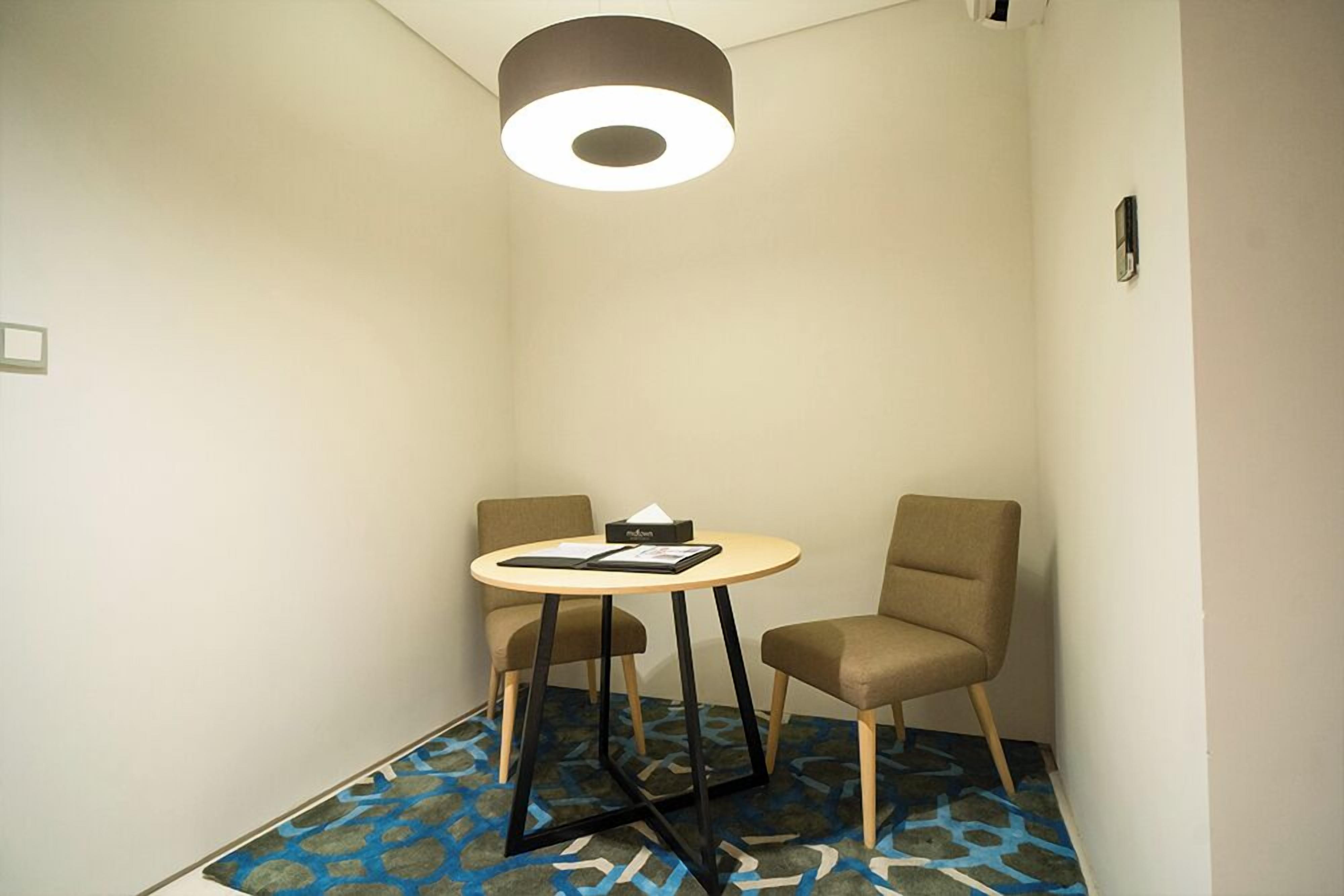 Quarto pequeno e moderno, com uma mesa redonda, duas cadeiras, tapete estampado e luminária no teto.
