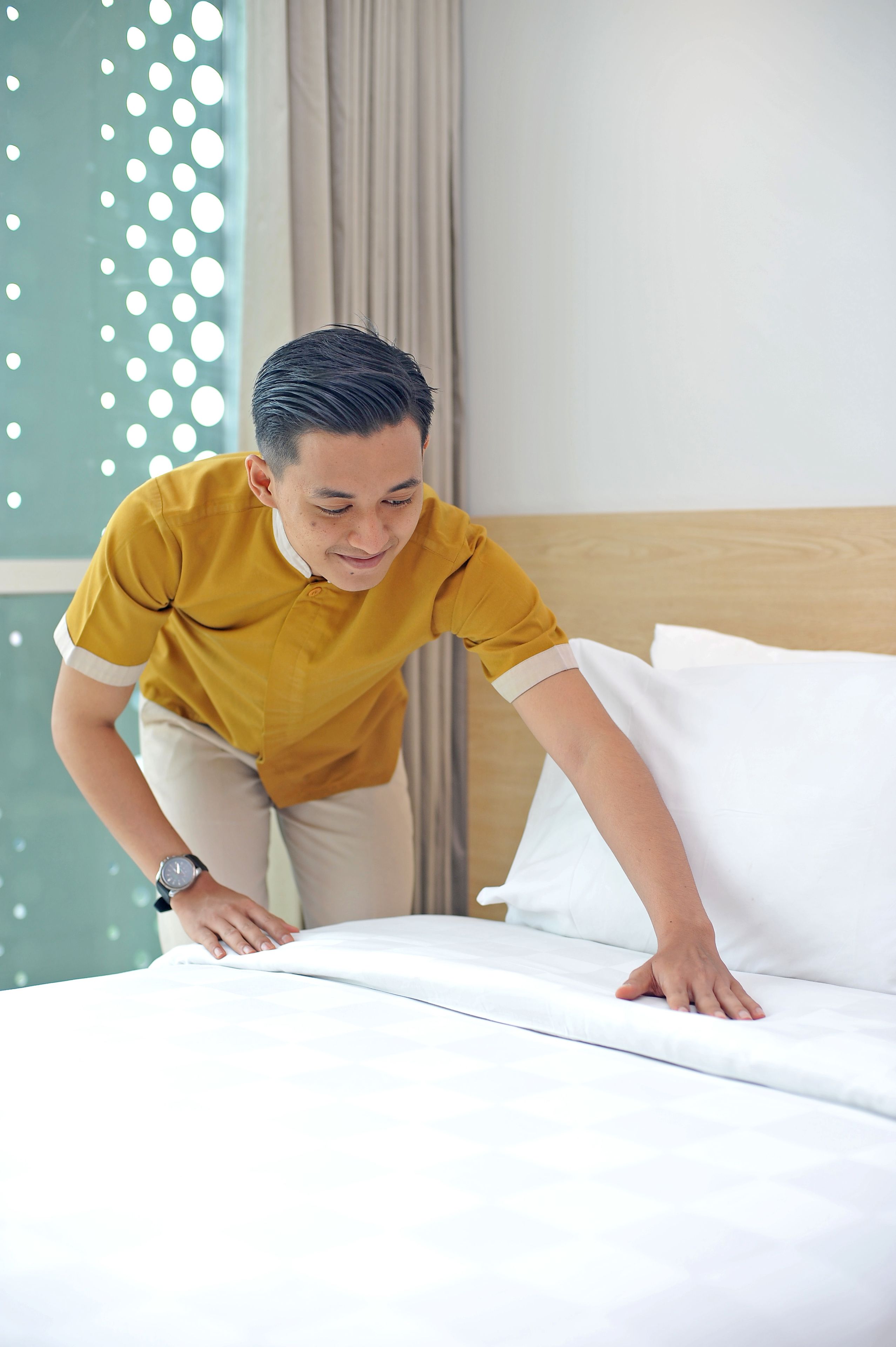 Iemand maakt een bed in een hotelkamer, met een houten hoofdbord en een groene muur met ronde patronen.
