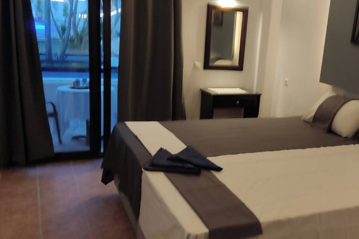 A hotel room with a bed, a small bedside table, a mirror, and a view of the outdoors.