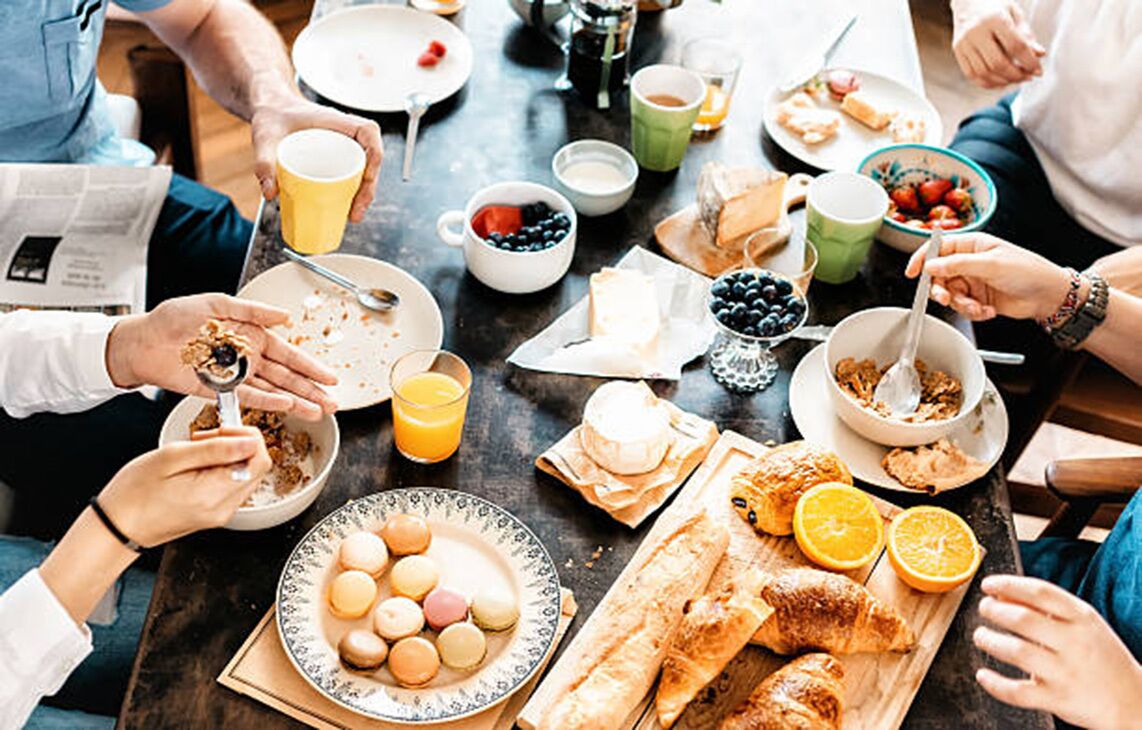 A group of people sitting around a table with breakfast items like eggs, bread, and fruit.