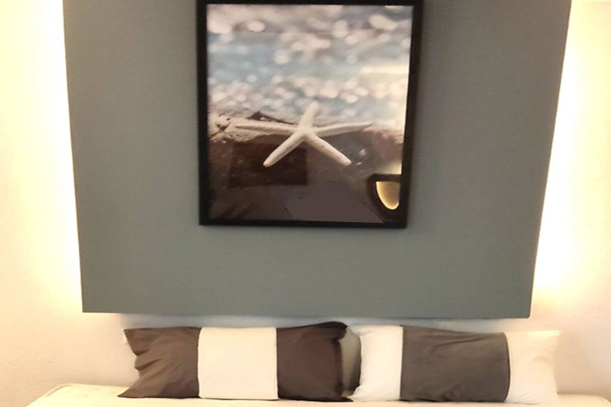 A bed with a white mattress, dark grey and white pillows, and a framed picture above.