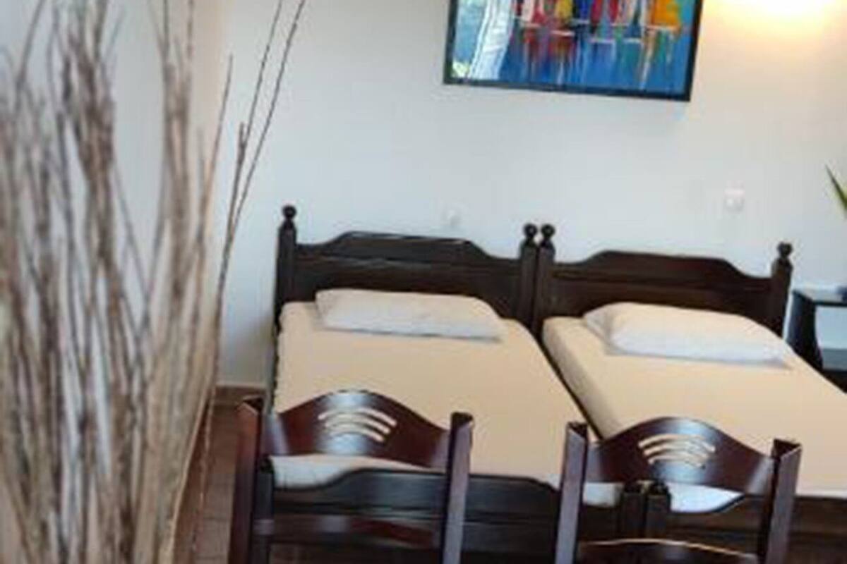 A room with two beds, a dining table with chairs, a painting on the wall, and a vase with branches.