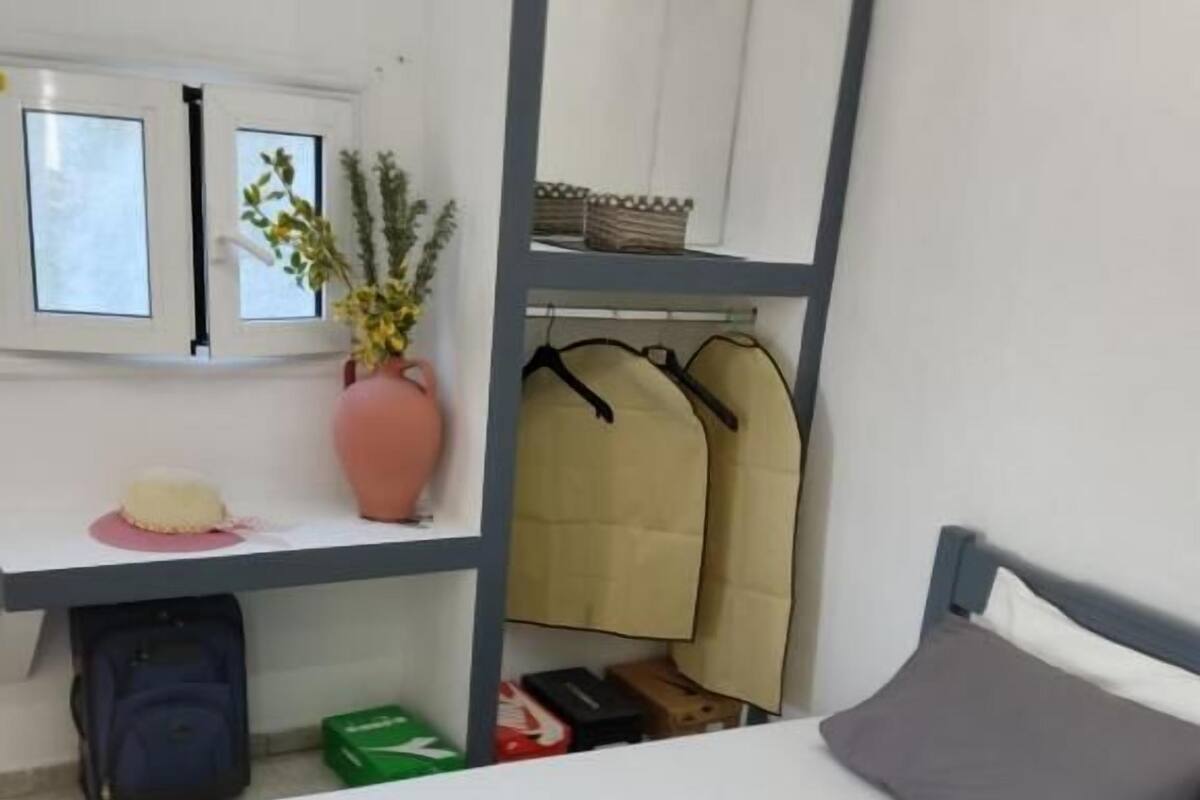 A single bed with a grey pillow and a white sheet, a small desk with a pink vase, a mirror, and a window with blinds.
