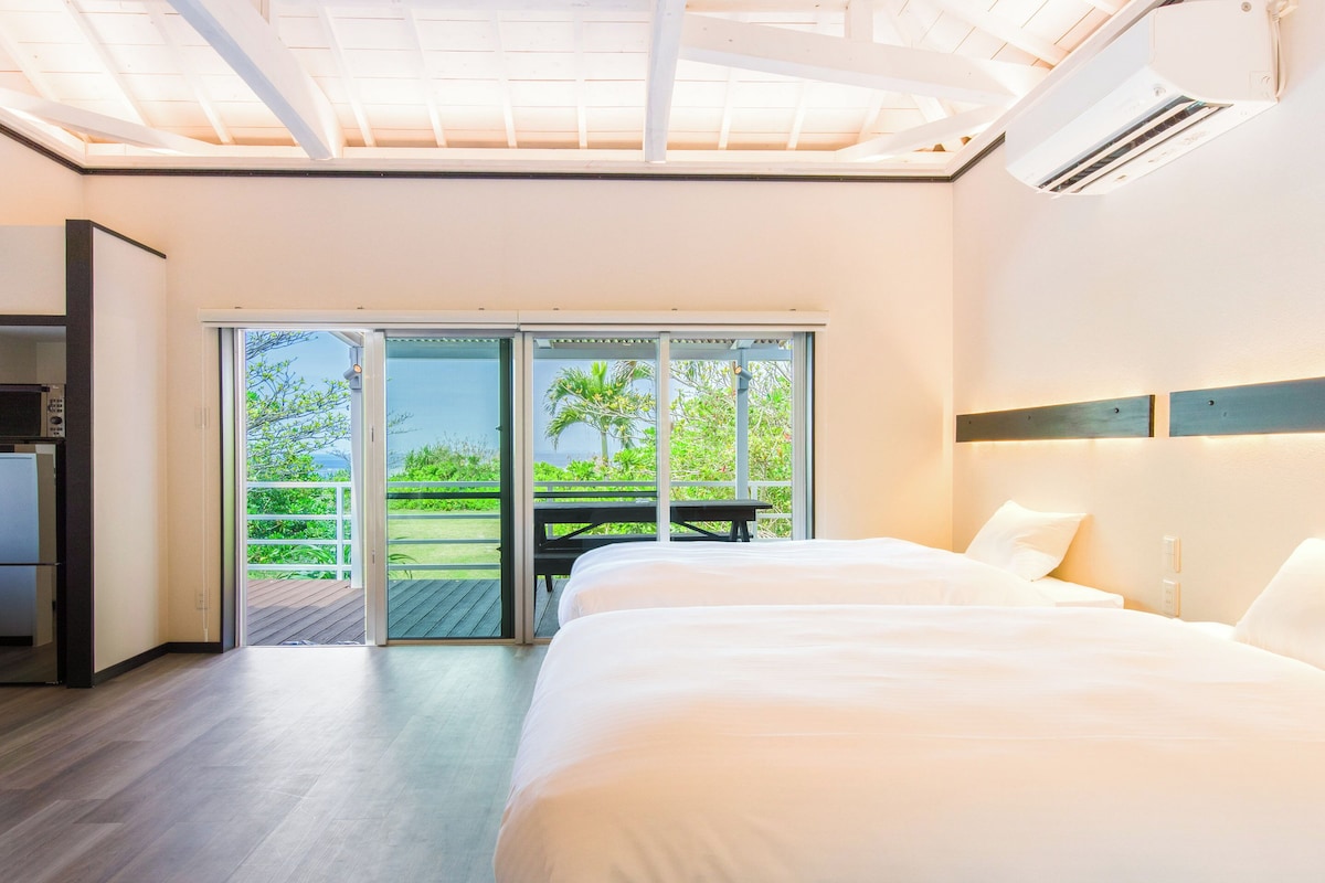 Een moderne hotelkamer met een groot bed, een bureau en een glazen schuifdeur naar een balkon met uitzicht op groen en de zee.