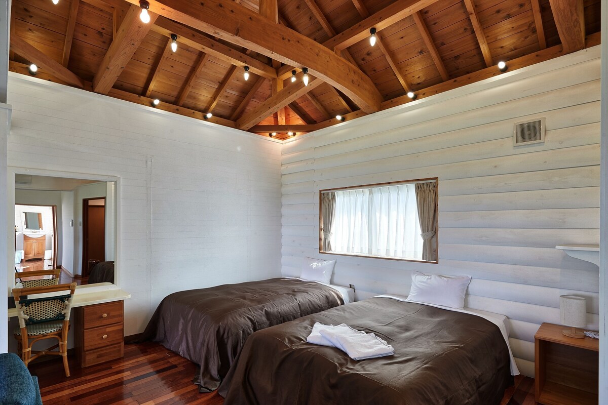 Een slaapkamer met twee bedden, houten balken in het plafond, een raam met gordijnen, een spiegel en een nachtkastje van hout.